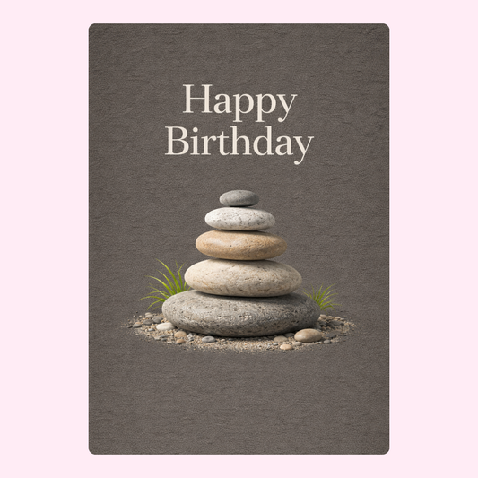 A tranquil birthday card featuring a stack of smooth stones arranged in a balanced formation with subtle grass accents, set against a muted gray background. The text "Happy Birthday" is elegantly displayed in white font.